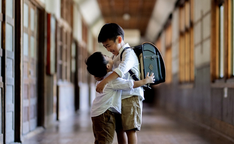 小学校の廊下。ランドセルを背負った兄と小さな弟が、仲良さそうに笑顔で抱きしめ合っている。