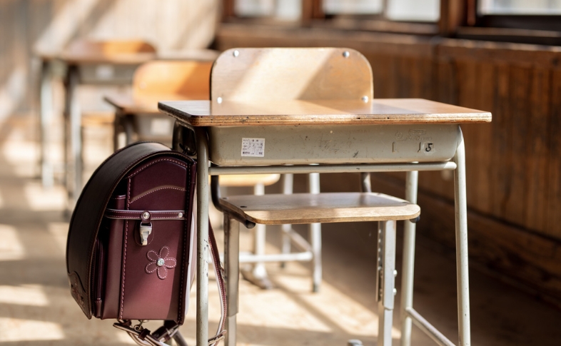 日が差し込む小学校の教室。学習机と椅子が並び、机には可愛いワイン色のランドセルが掛けられている。